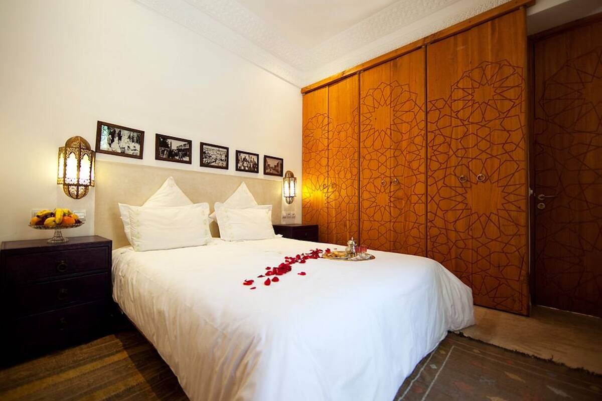 A neatly made bed with white linens, a bedside table with a fruit bowl, and a wall adorned with framed pictures.