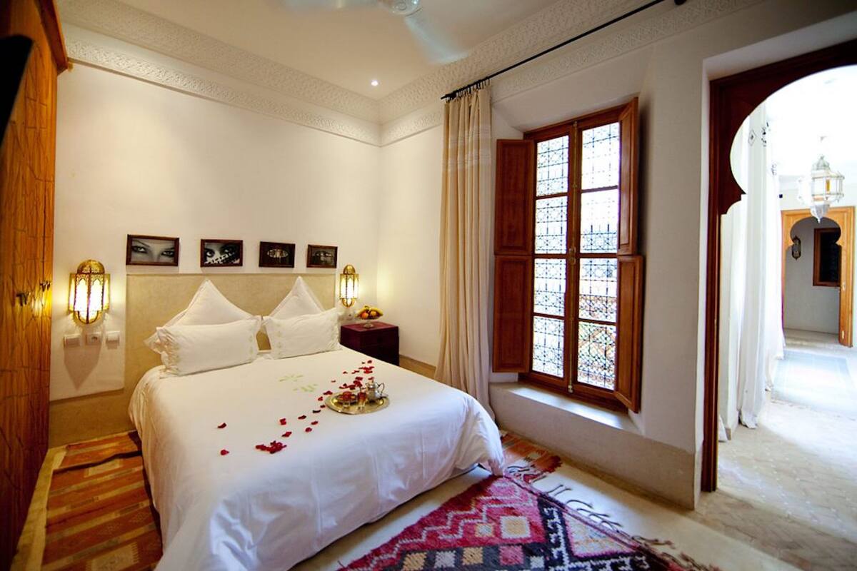 A hotel room with a large bed, a nightstand with a lamp, a window with curtains, and a view of the outside.