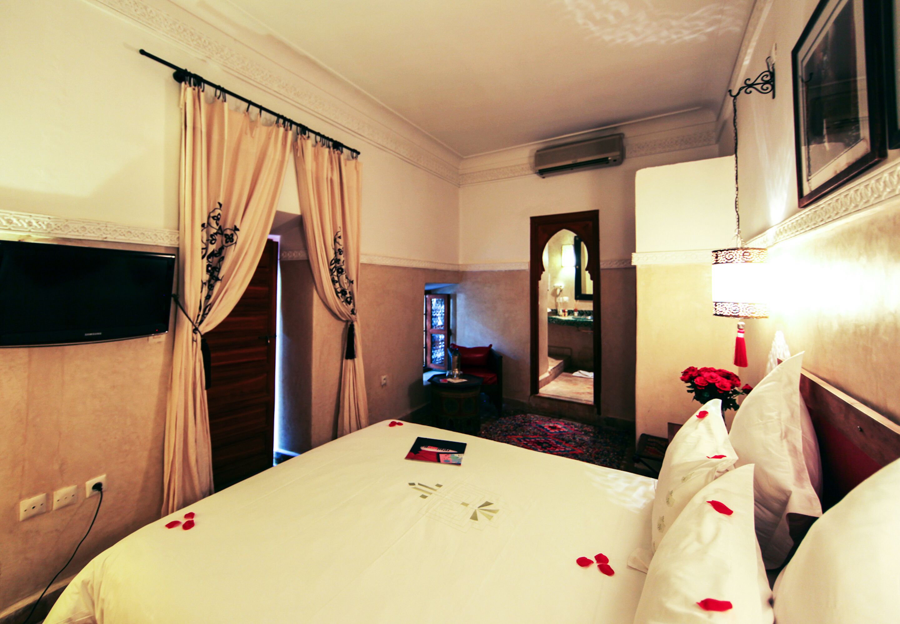 A hotel room with a large bed, a flat-screen TV mounted on the wall, a bedside table with a lamp, and a mirror above the bed.