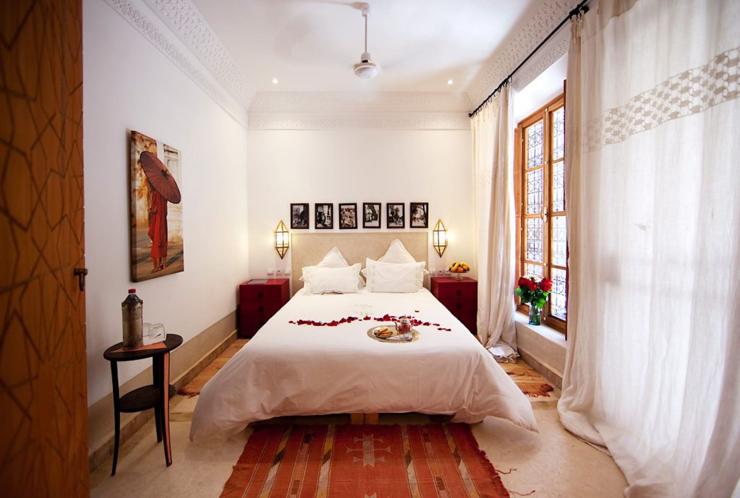Une chambre à coucher bien rangée, avec un grand lit, deux tables de chevet, une fenêtre avec des rideaux et des tableaux encadrés accrochés au mur.