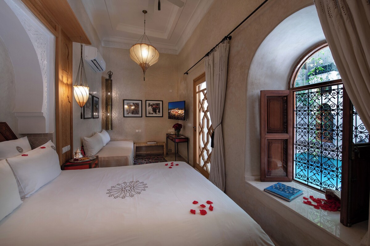 A hotel room with a large bed, a sitting area with a sofa, a small table with a vase of flowers, and a window with decorative ironwork.