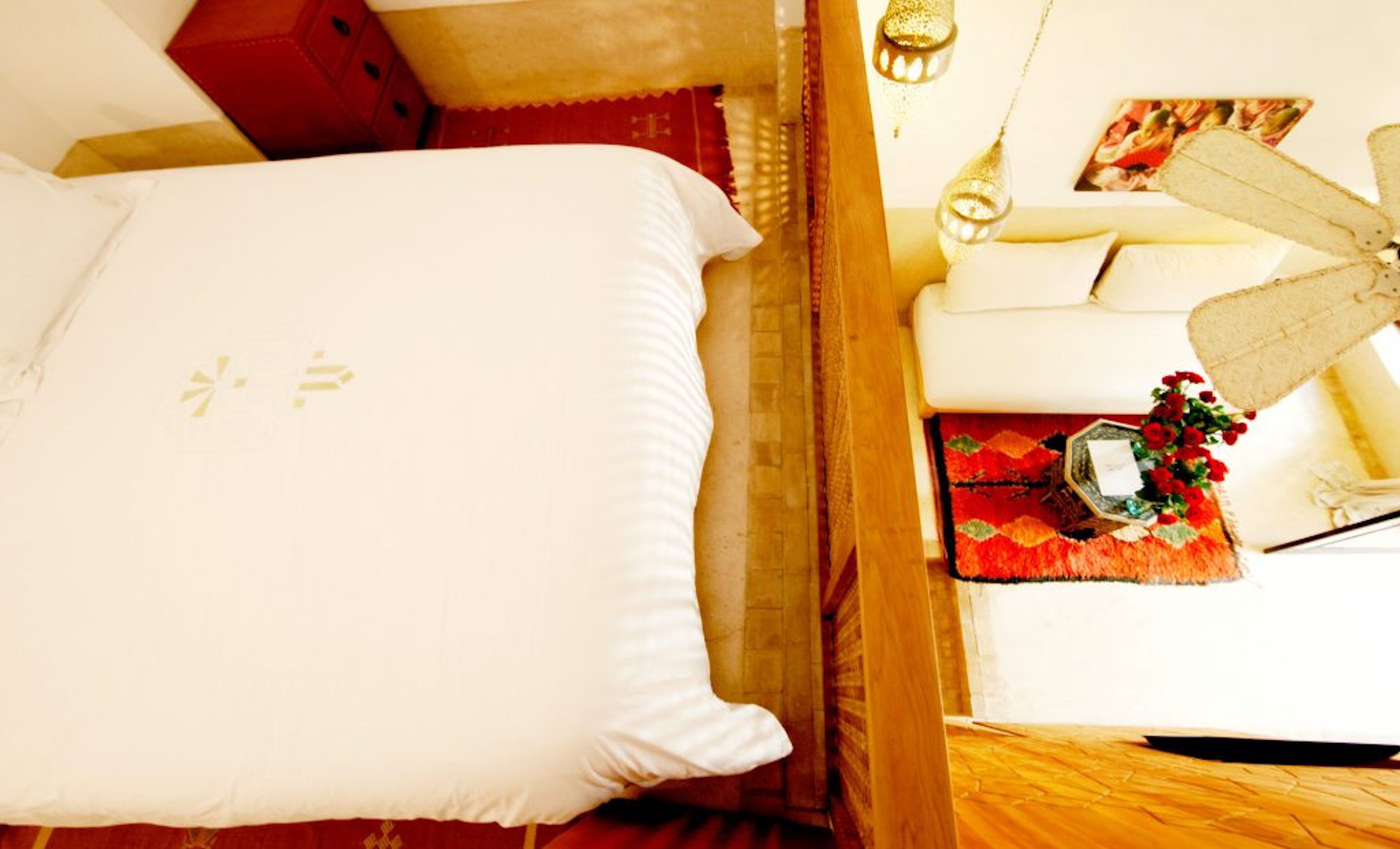 A bed with a white pillow and a red headboard, a wooden floor, and a small table with a red rug and a vase of red flowers.