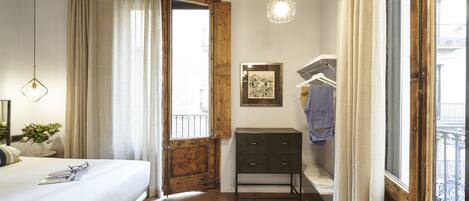 Un dormitorio con suelo de madera, una cama con colcha blanca, una mesita de noche con lámpara, un armario de madera y una ventana con vista a un balcón.