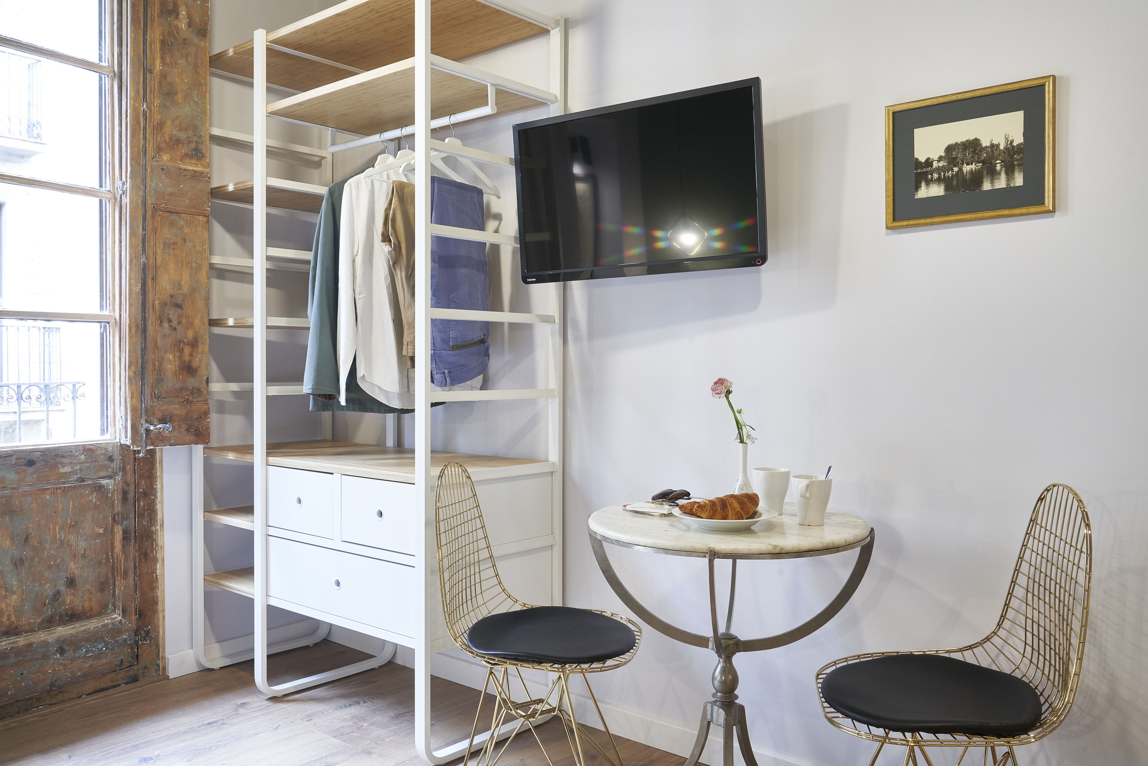 Een kamer met een witte kast, een aan de muur bevestigde televisie, een klein rond tafeltje met twee stoelen en een raam met uitzicht op gebouwen.
