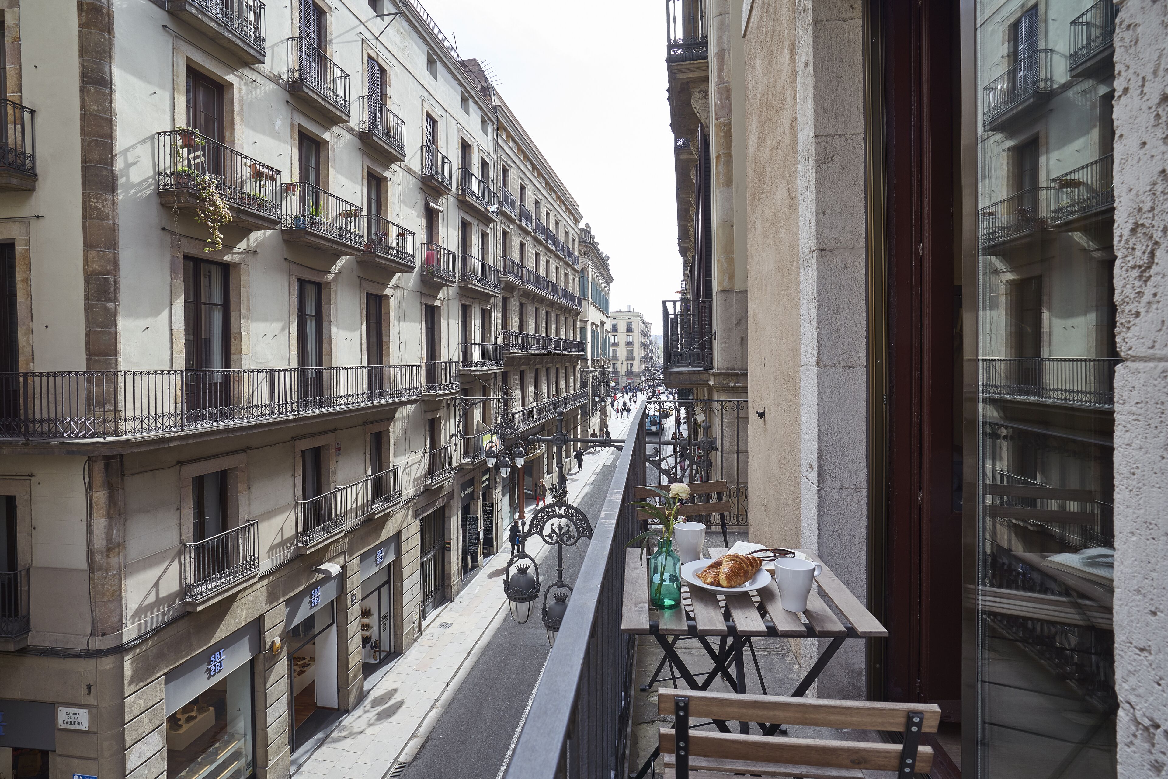 Une rue étroite, bordée de bâtiments des deux côtés, avec des balcons ornés de plantes et des espaces aménagés pour s’asseoir à l’extérieur.