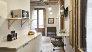 A kitchen with a microwave, toaster, and fruit bowl on the counter, wooden beams on the ceiling, and a dining area with chairs and a table. - AinB Gothic-Jaume I Apartments (Barcelona)