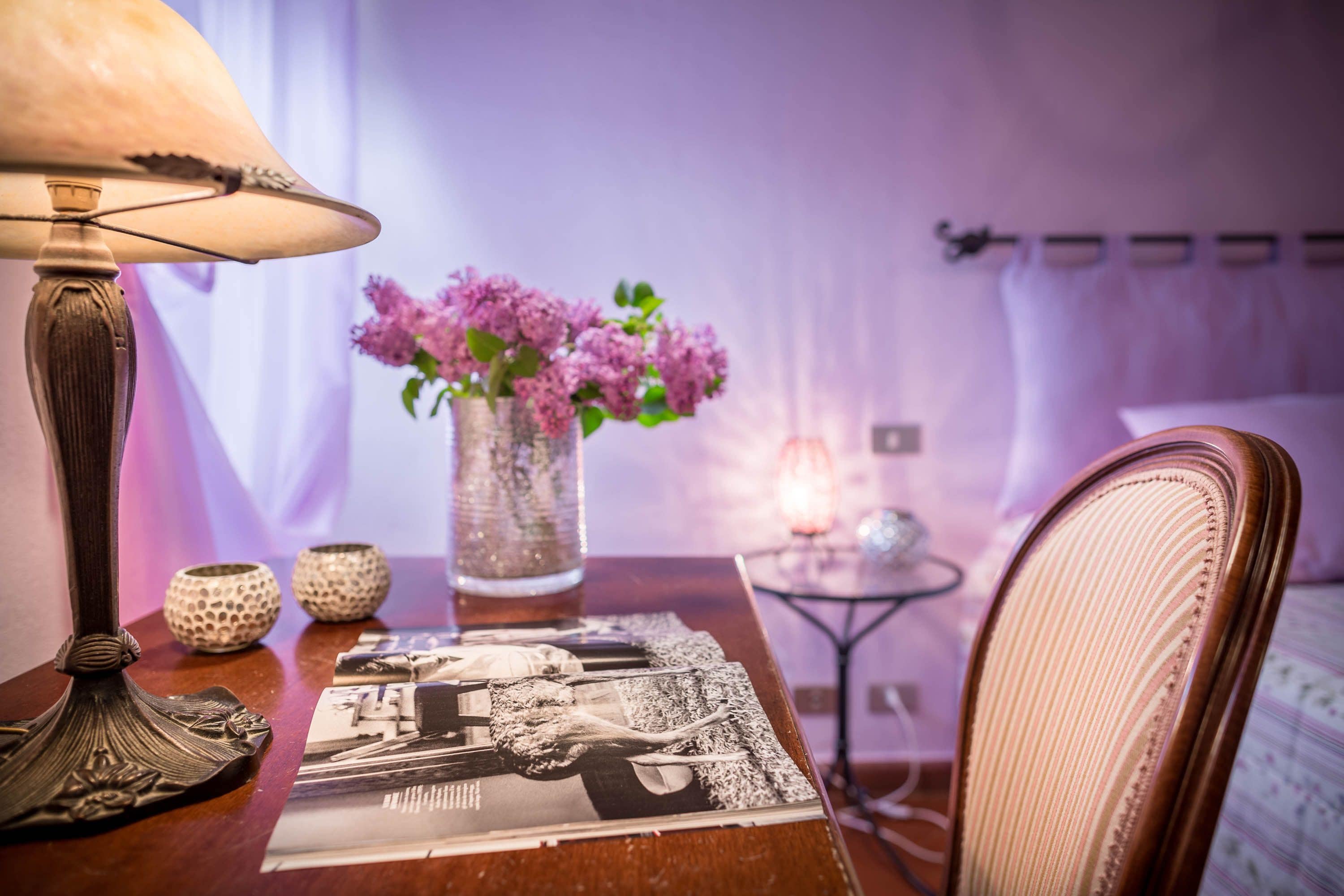 A neatly arranged room with a table featuring a lamp, a vase with flowers, and a magazine. A chair is positioned near the table, and a bed with a white headboard is visible in the background.