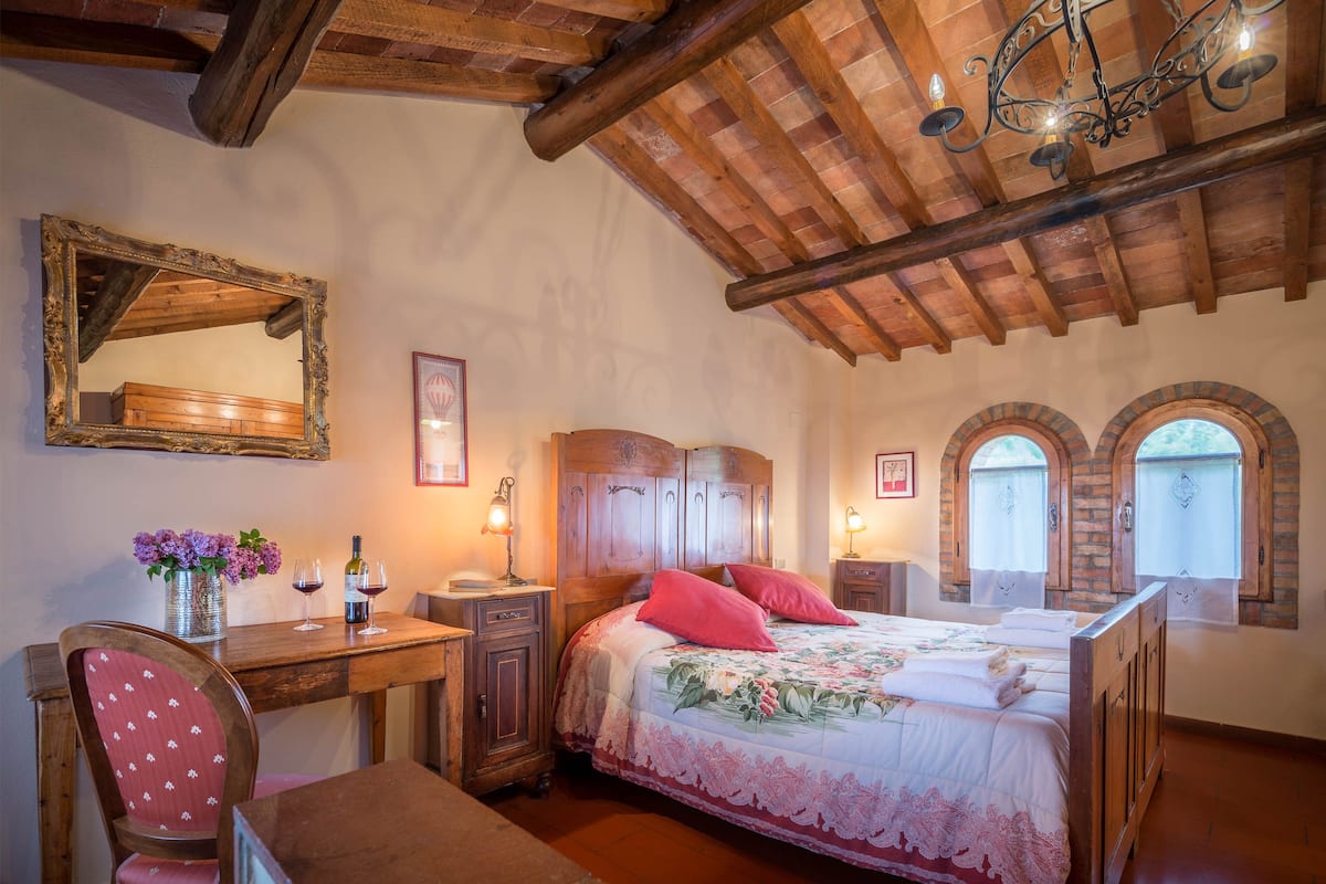 Une chambre avec un plafond en bois, un lit recouvert d’une literie à motifs floraux, un bureau avec une chaise et un miroir accroché au mur.
