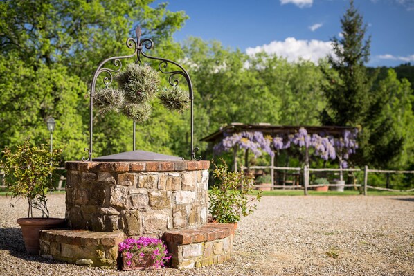 A stone structure with a metal arch, surrounded by gravel, potted plants, and a wooden fence with purple flowers.