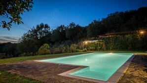 Una piscina rectangular con cubierta de piedra y borde de madera, rodeada de césped y árboles.