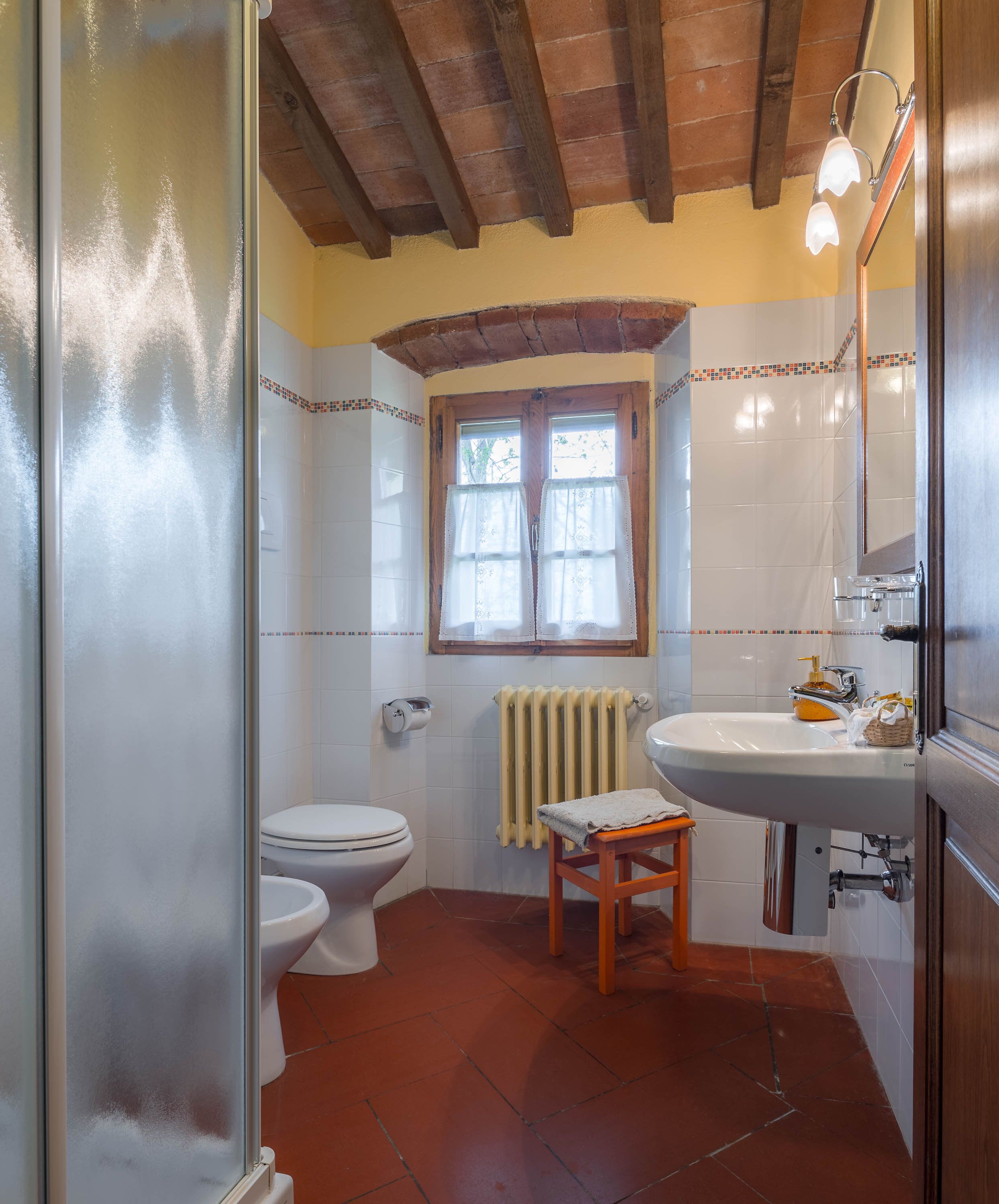A bathroom with a toilet, a sink, a window with curtains, and a wooden stool.