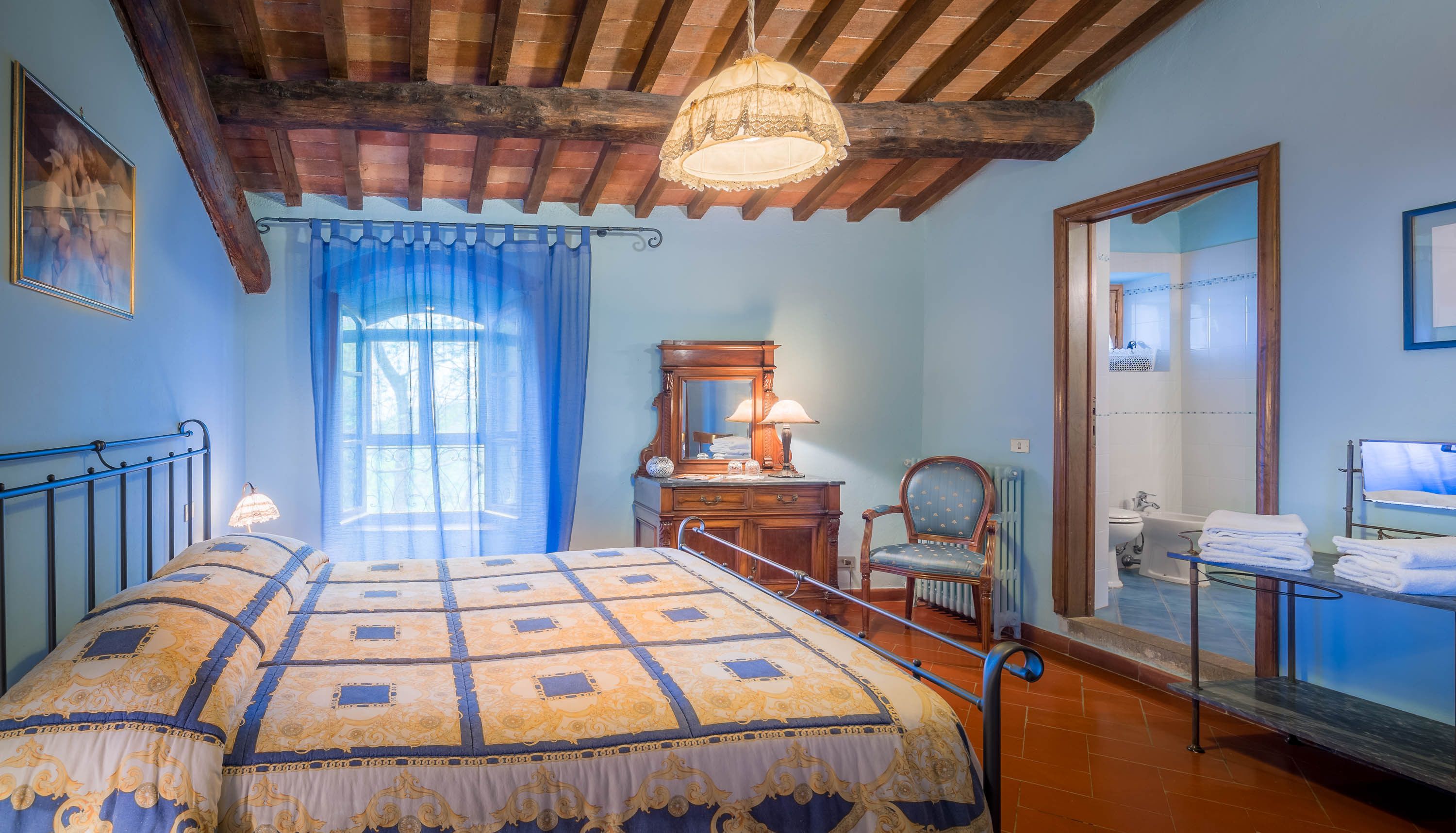 Quarto com cama, uma cadeira, cômoda, espelho e banheiro visível através de uma porta aberta.
