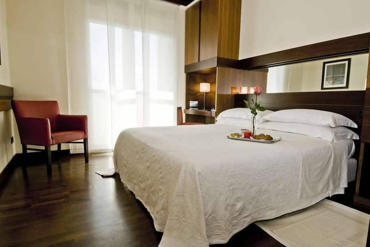 A neatly made bed with a tray of food and a rose, a red chair, and a bedside table with a lamp.