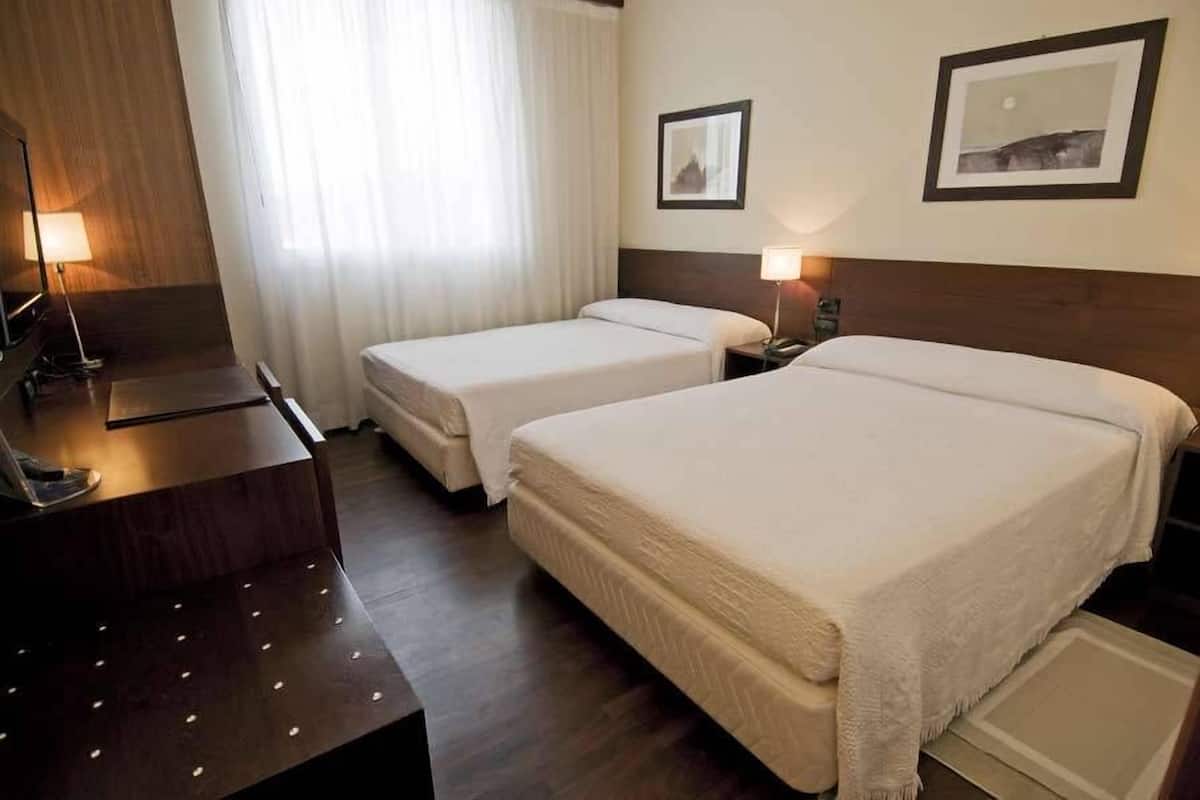 A hotel room with two single beds, a desk with a lamp, a nightstand with a phone, and a window with curtains.