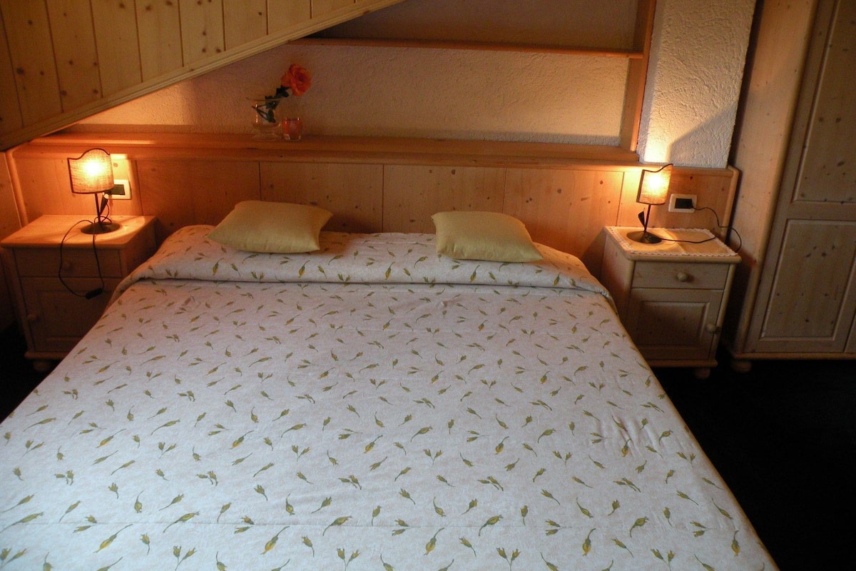 A bedroom with a bed, two bedside tables, a nightstand, a lamp, and a small vase with a flower.