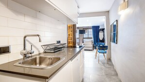 A modern kitchen with a stainless steel sink, white tiled backsplash, and a wooden door leading to a living area with a sofa and blue chairs. - AinB Picasso Corders Apartments (Barcelona)