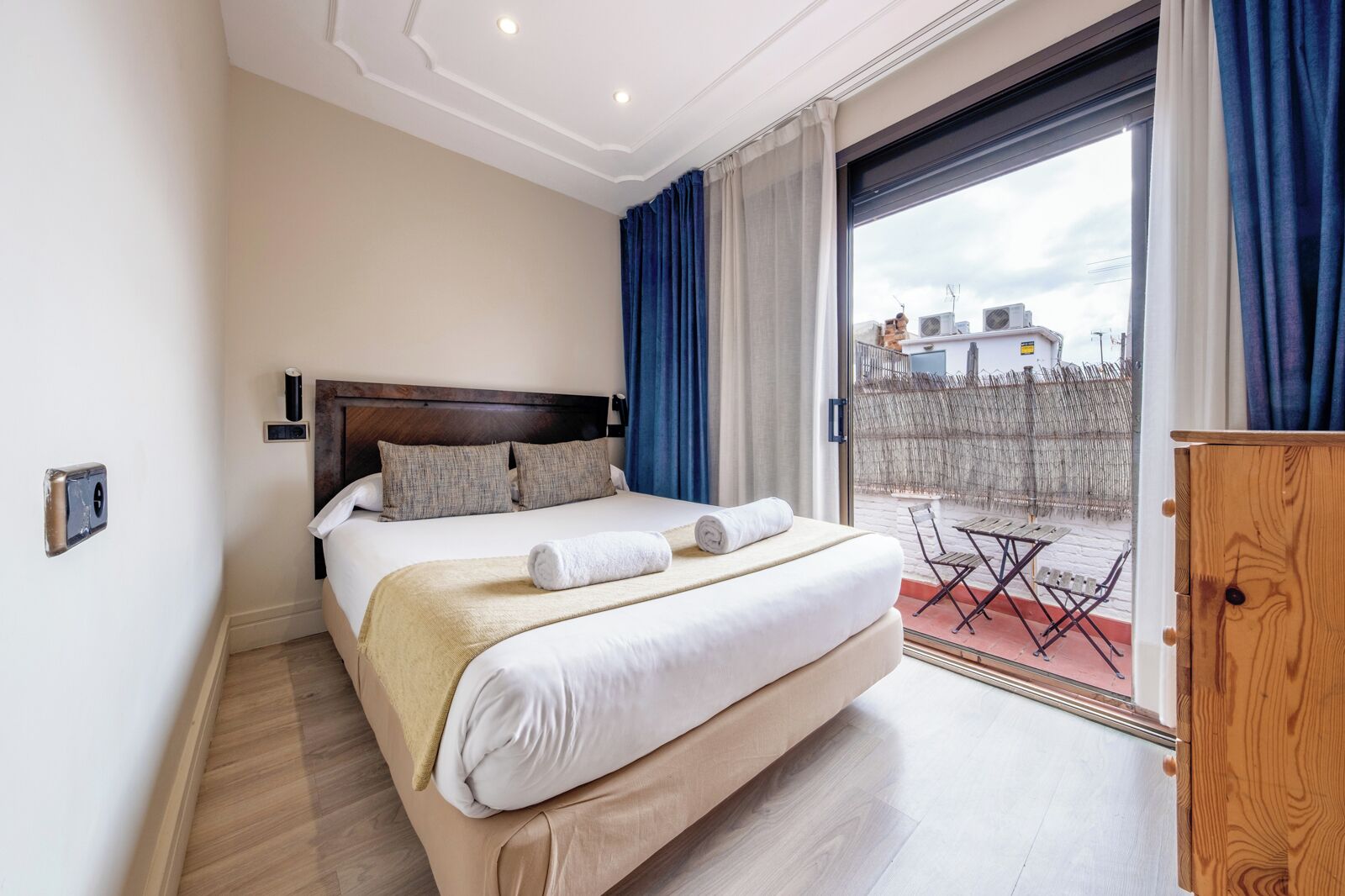 Habitación de hotel con una cama grande, cabecera de madera, balcón con vistas a edificios y un armario de madera.