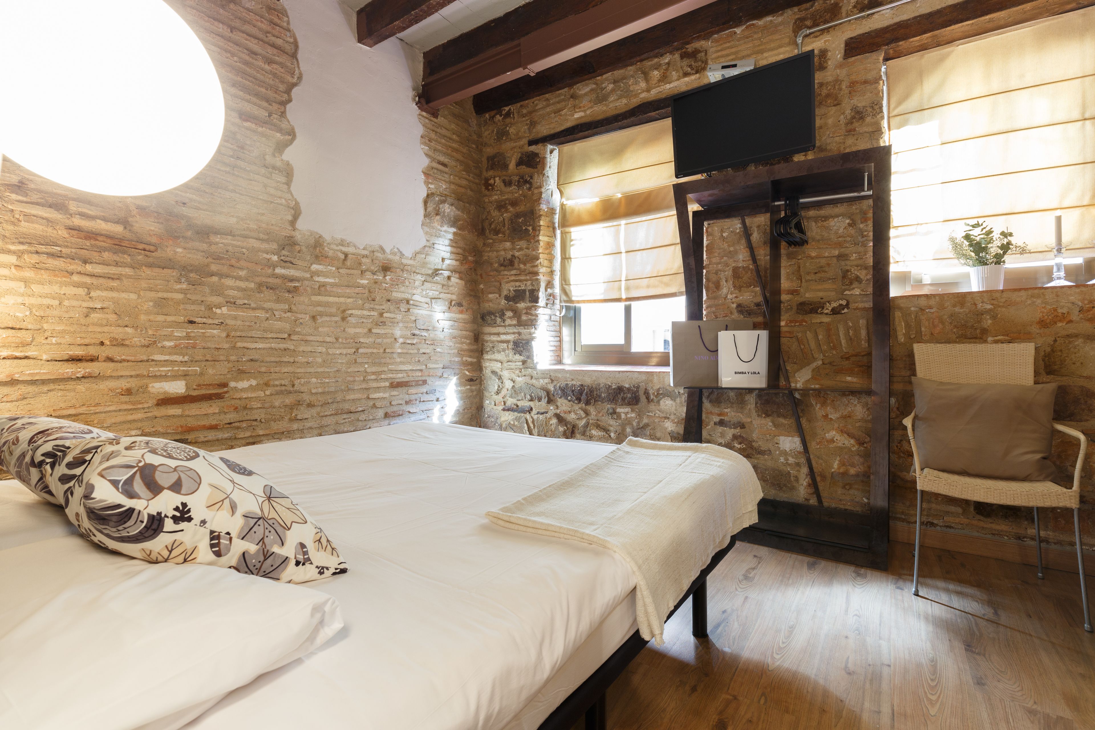 A room with a bed, a chair, a window with curtains, and a stone wall.