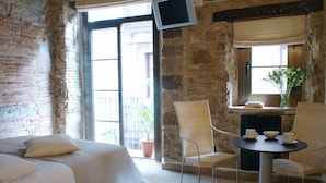 A room with a brick wall, a bed with white bedding, two wicker chairs, a small round table with a bowl and cups, and a TV mounted on the wall. - AinB Picasso Corders Apartments (Barcelona)