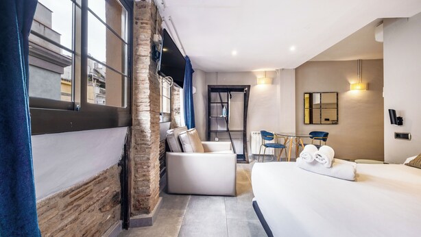 A modern hotel room with a large bed, a seating area with a sofa and chairs, a small table, and a view through the window. - AinB Picasso Corders Apartments (Barcelona)