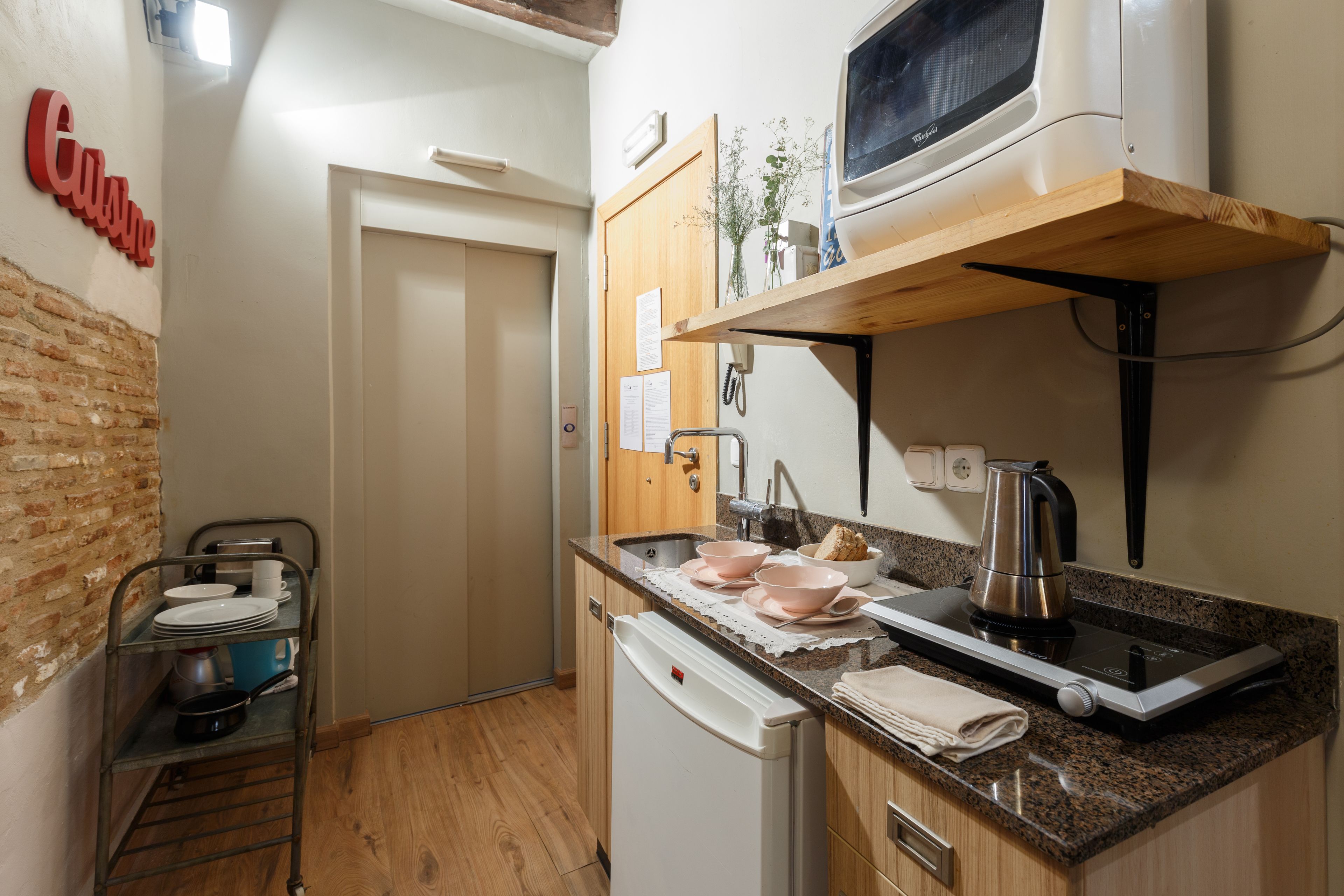 Een kleine keuken met een magnetron, waterkoker en een vaatwasser. Een metalen plank met servies en een deur naar een andere kamer.