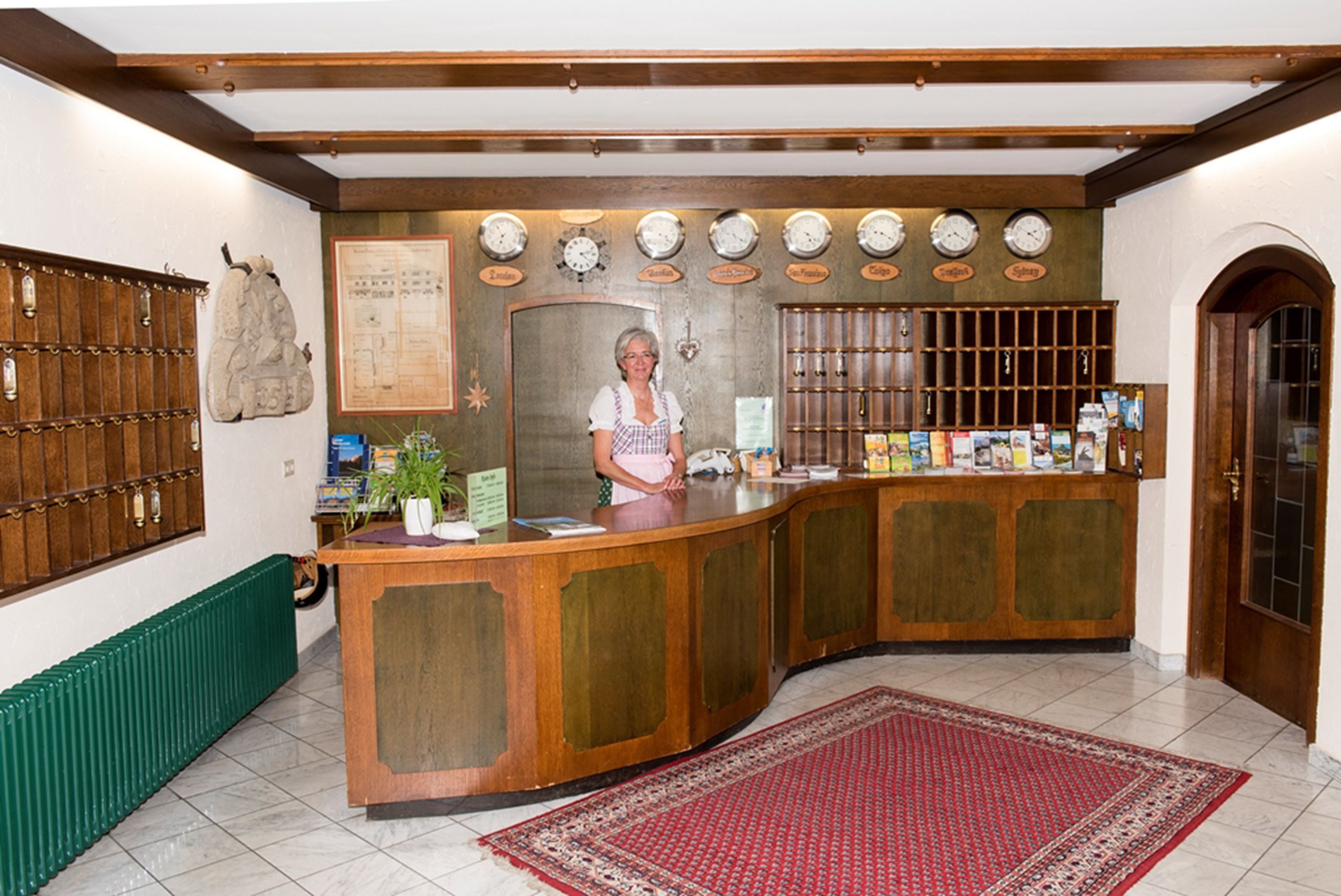Un área de recepción con un mostrador de madera, una mujer de pie detrás y una pared con relojes y un mapa.