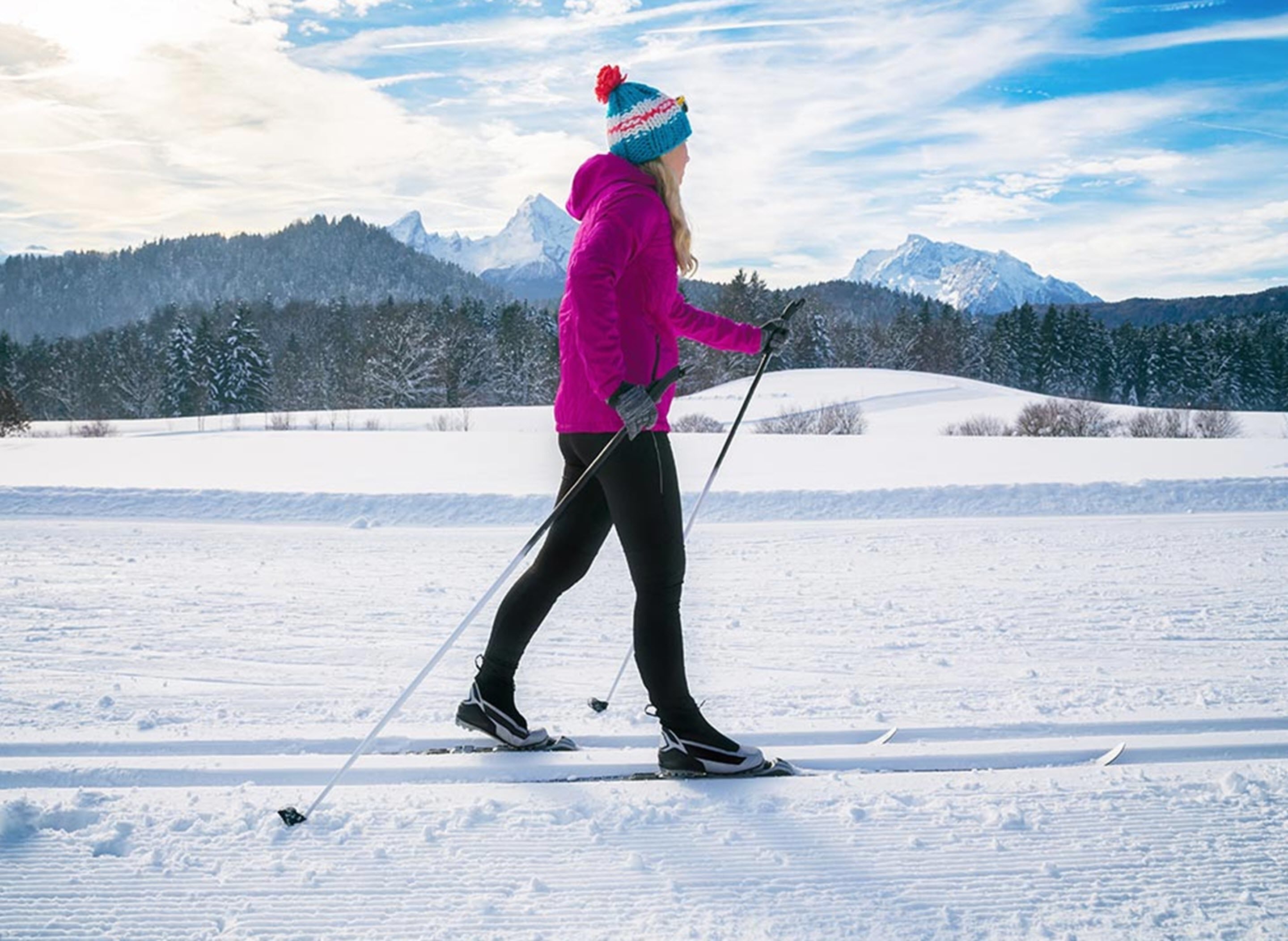Eine Person langlauft in einer verschneiten Landschaft mit Bergen im Hintergrund.