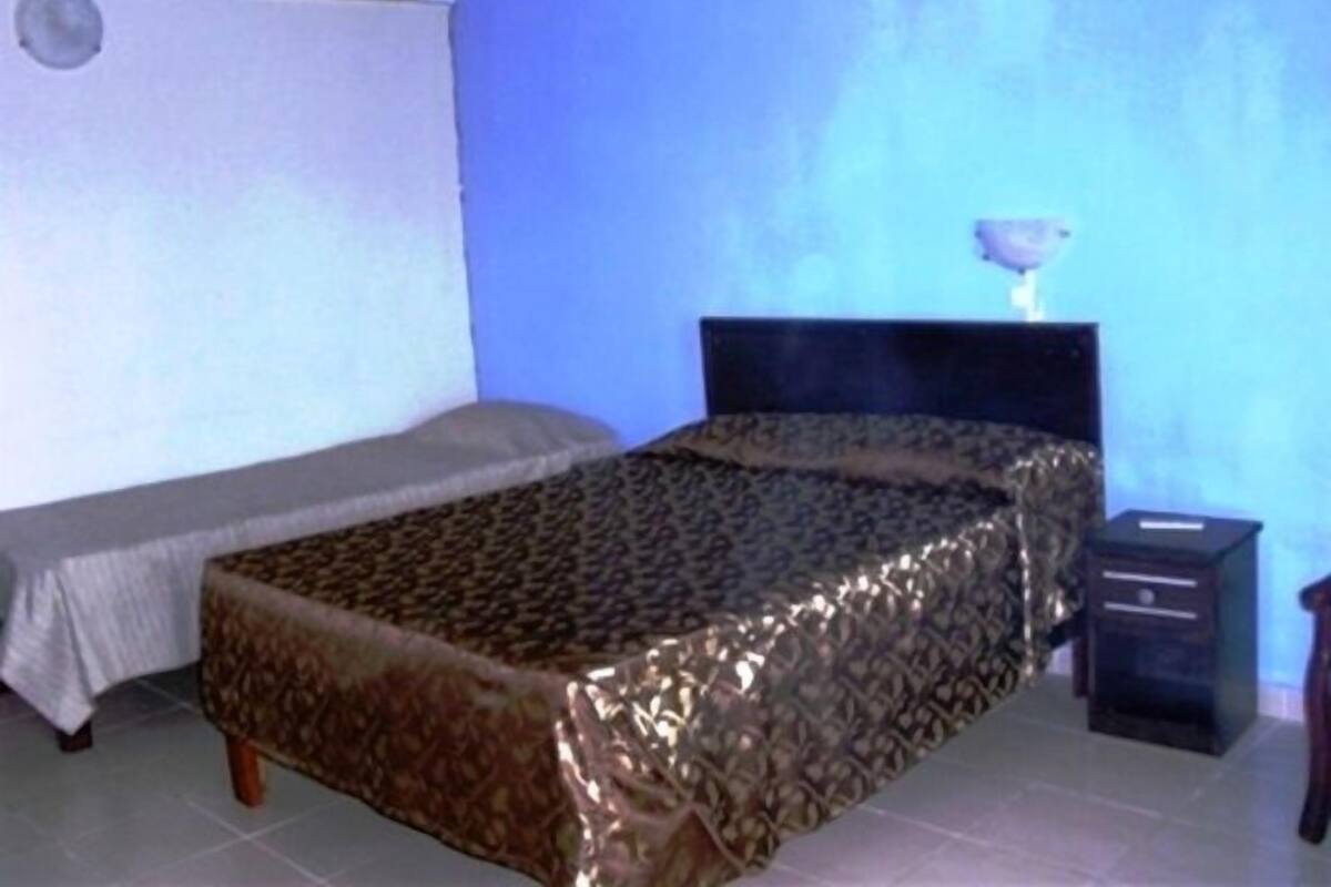 A small, single-bed room with a patterned bedspread, a bedside table, and a wall-mounted air conditioning unit.