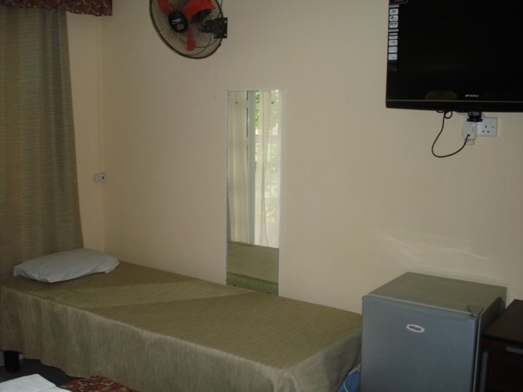 A single bed with a headboard, a small desk with a computer, a wall-mounted TV, and a window with curtains.
