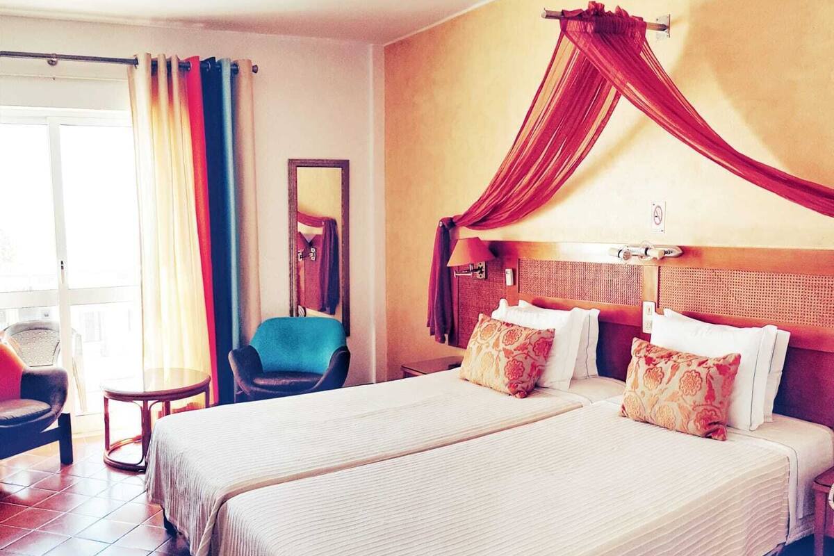 Een hotelkamer met twee bedden, een stoel, een raam met gordijnen en een balkon met een rode luifel.