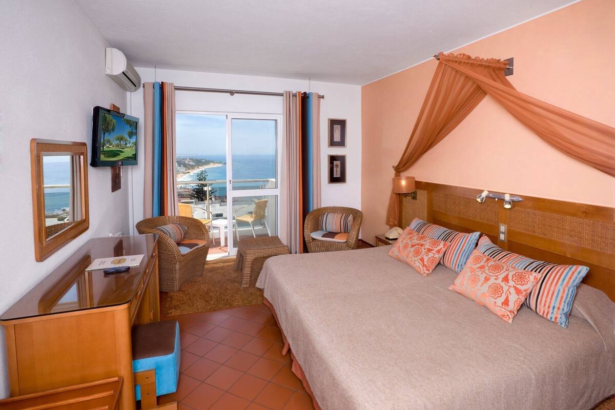 Een hotelkamer met een bed, een bureau, een stoel, een kleine tafel en uitzicht op de oceaan door een glazen schuifdeur.