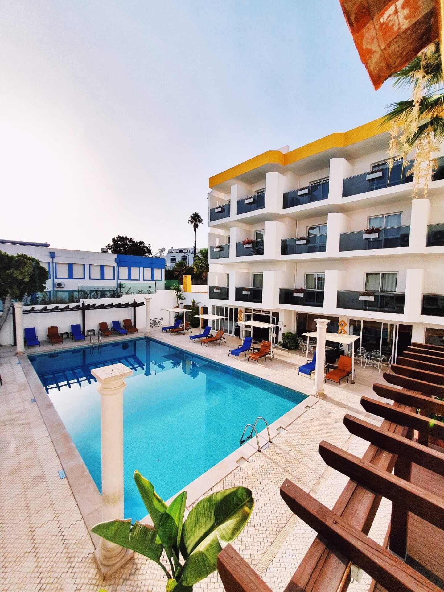 A hotel with a swimming pool, outdoor seating, and a multi-story building with balconies.