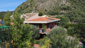 Una casa con techo rojo, rodeada de follaje y una colina rocosa al fondo.