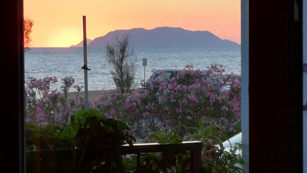 Una vista del atardecer sobre el agua, con montañas al fondo, un balcón con plantas y una valla.