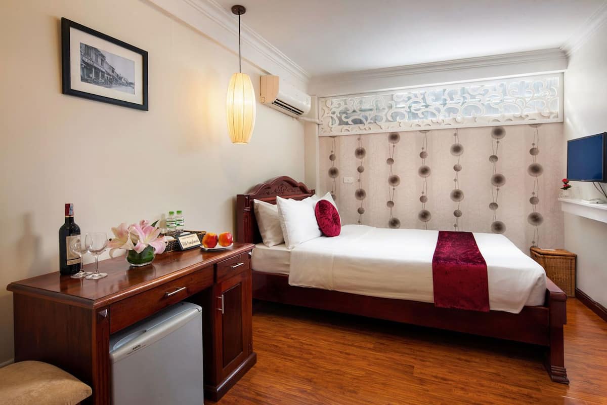 Habitació d'hotel amb llit, escriptori amb ordinador, nevera petita i finestra amb cortina decorativa.