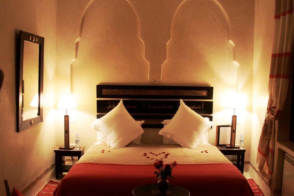A hotel room with a large bed, two bedside tables, a chair, a small table with a vase, and a mirror on the wall.