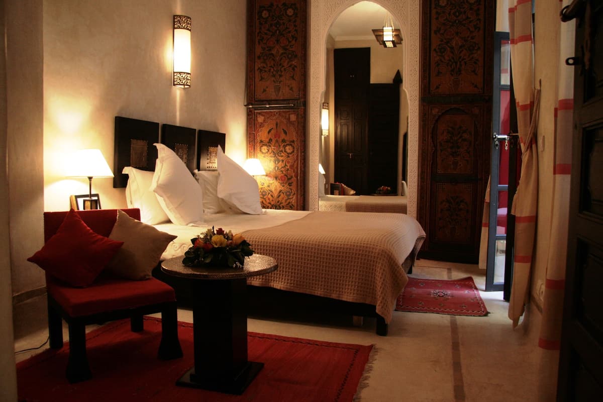 A hotel room with a bed, two bedside lamps, a red chair, a small table with a vase of flowers, and a red rug.
