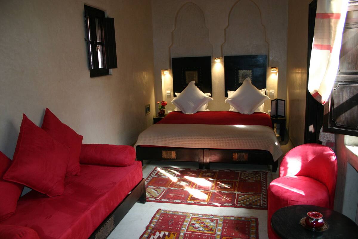 A hotel room with a large bed, red sofa, and a small table with a red candle.