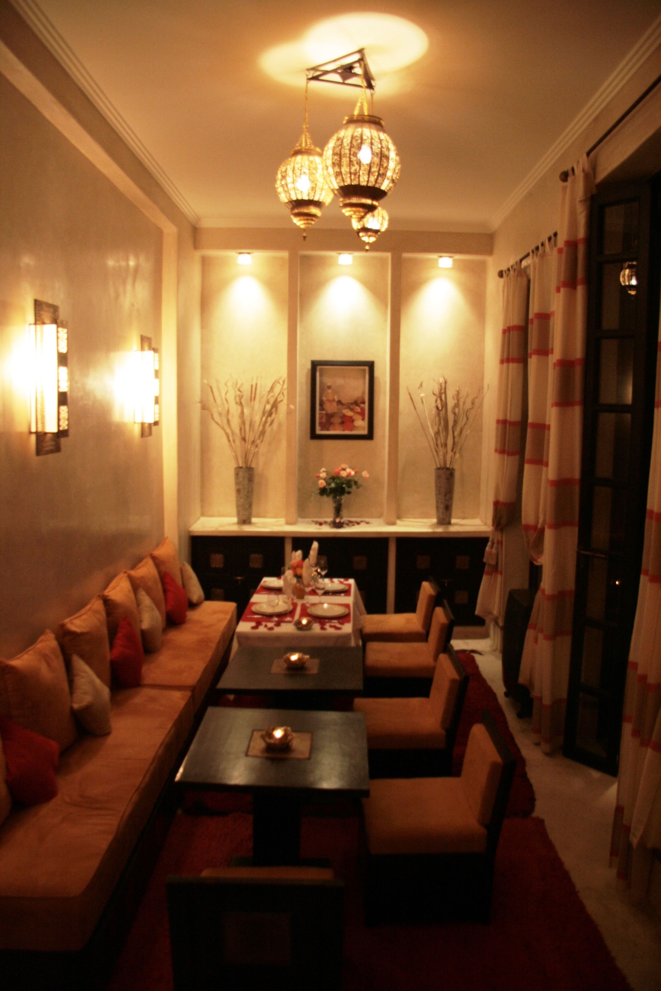 A dimly lit room with a sofa, two tables, and chairs arranged around a central table. There are wall sconces, a chandelier, and two vases with branches.
