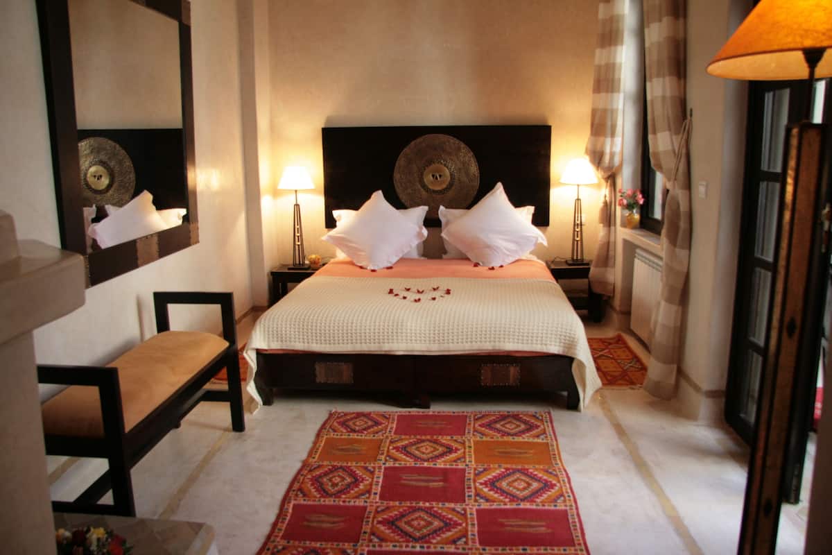 A hotel room with a large bed, two bedside lamps, a mirror, a bench, and a patterned rug.
