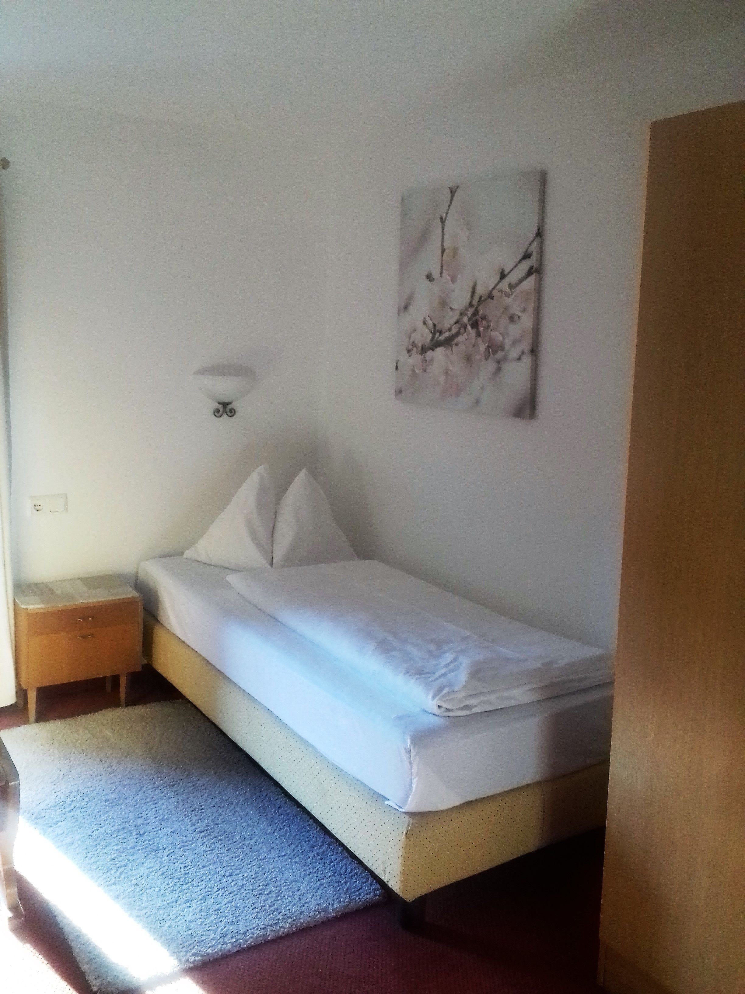 Ein Einzelbett mit weißer Matratze und Kissen, ein kleiner Nachttisch mit Schubladen, eine Wandleuchte sowie ein Bilderrahmen mit einem Zweig voller rosa Blüten.