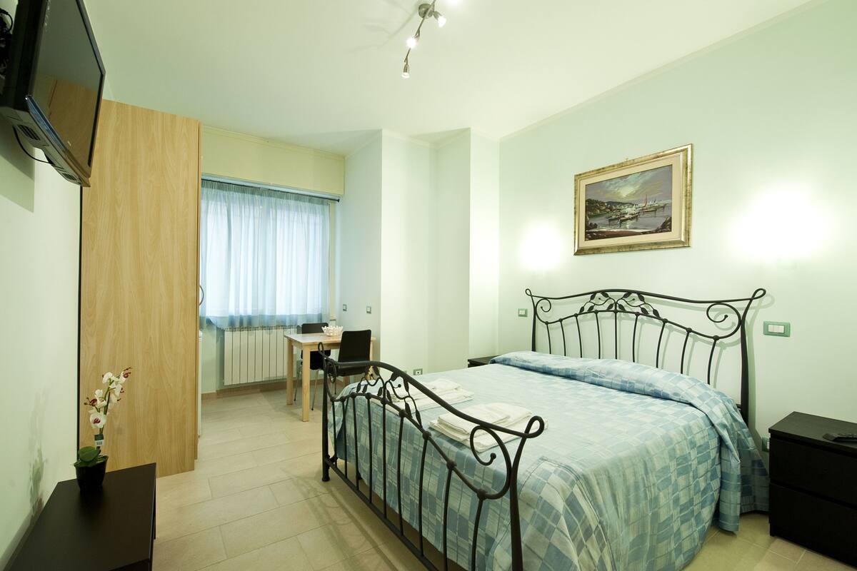 A bedroom with a bed, a desk, a chair, a TV, and a painting on the wall.