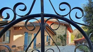 Una reja de hierro forjado con un diseño decorativo, que da acceso a un edificio de fachada de piedra y contraventanas azules.