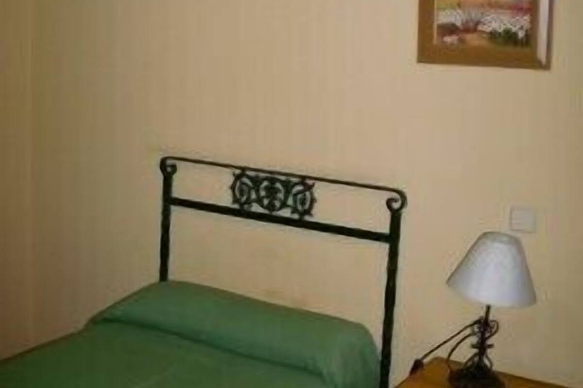 A single bed with a green bedspread, a wooden nightstand with a lamp, and a framed picture on the wall.