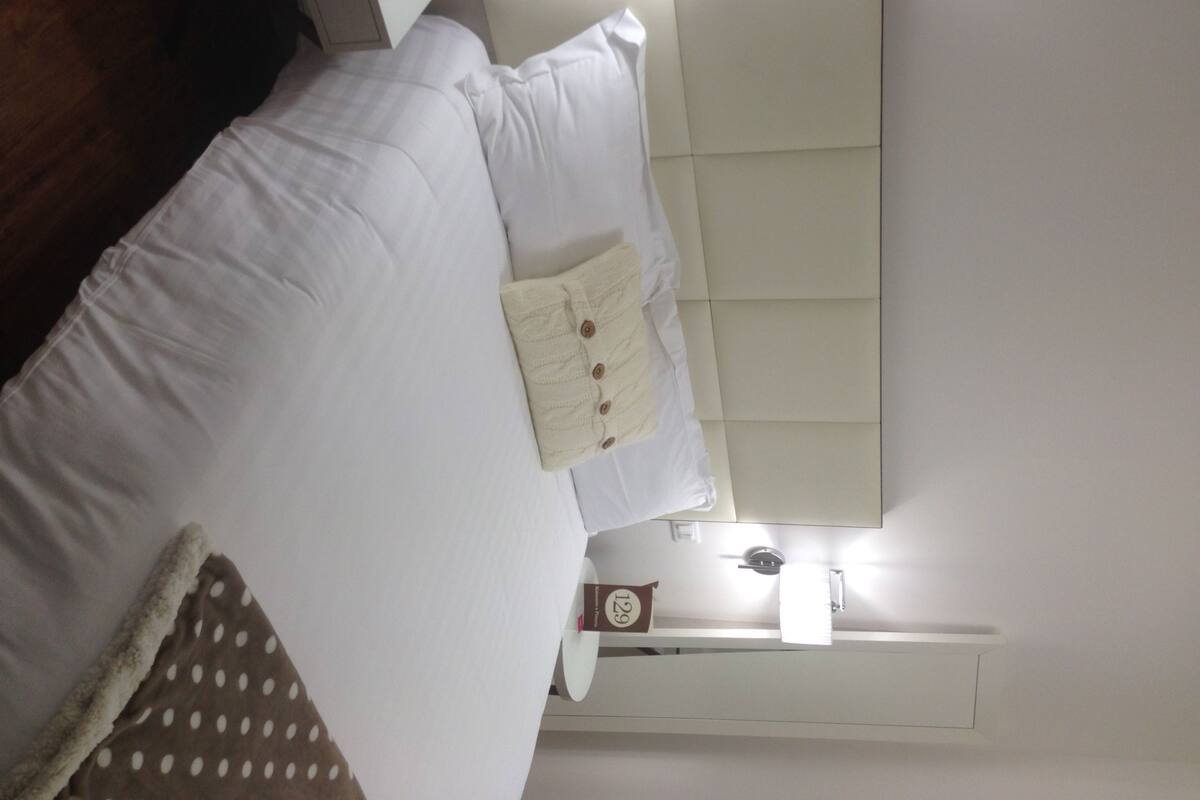 A hotel room with a bed, a white headboard, a white pillow, a white blanket, and a white polka dot blanket.