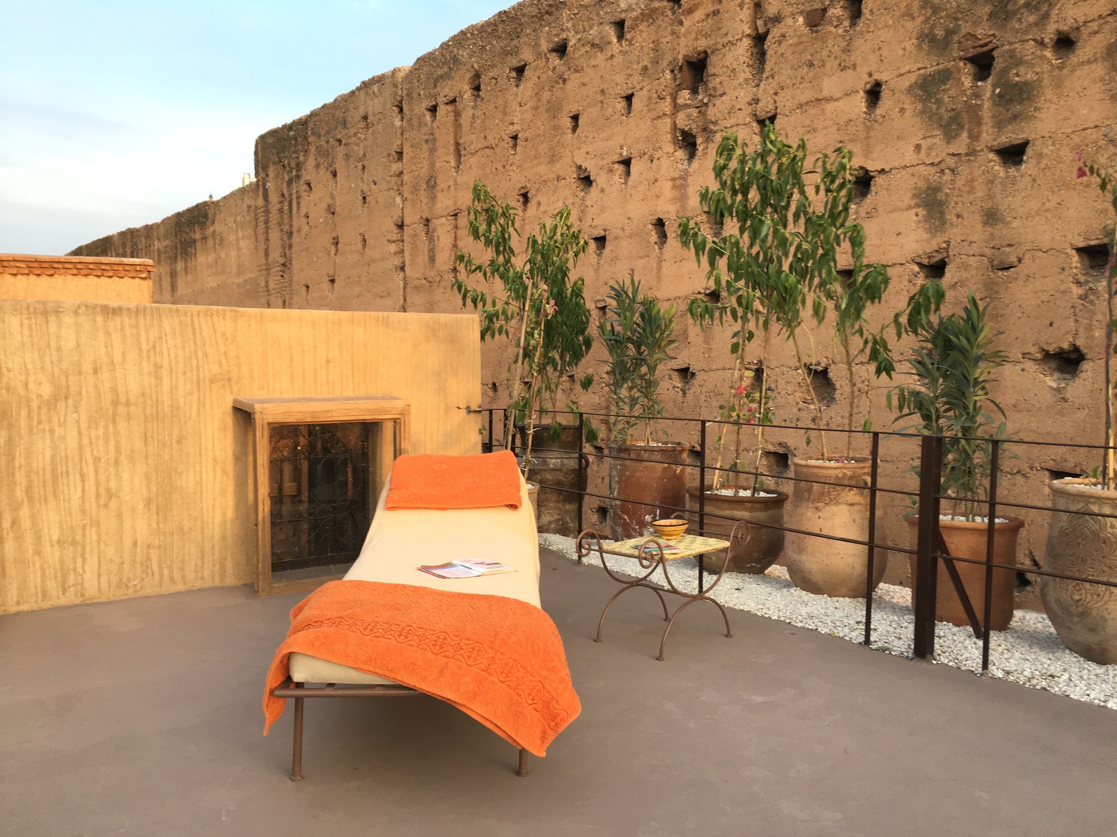 A single bed with an orange blanket on a rooftop terrace with a view of a brick wall and potted plants.