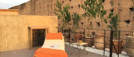 A single bed with an orange blanket on a rooftop terrace with a view of a brick wall and potted plants.