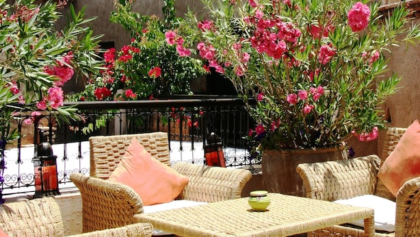 A patio with wicker furniture, a table, and potted plants with pink flowers. - Hotel Riad Carina (Marrakech)