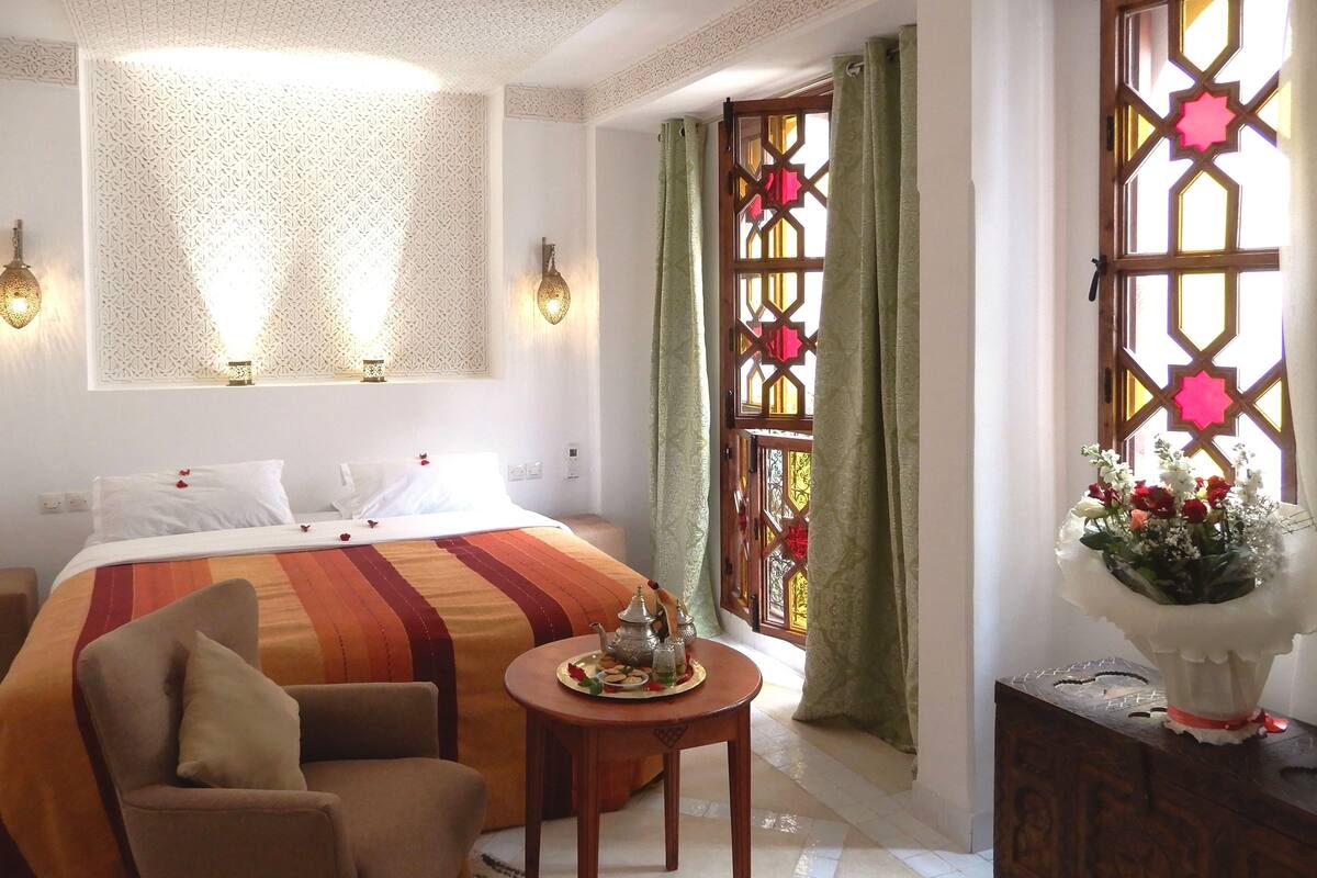 A hotel room with a bed, a chair, a small table with a tray, and a decorative vase.