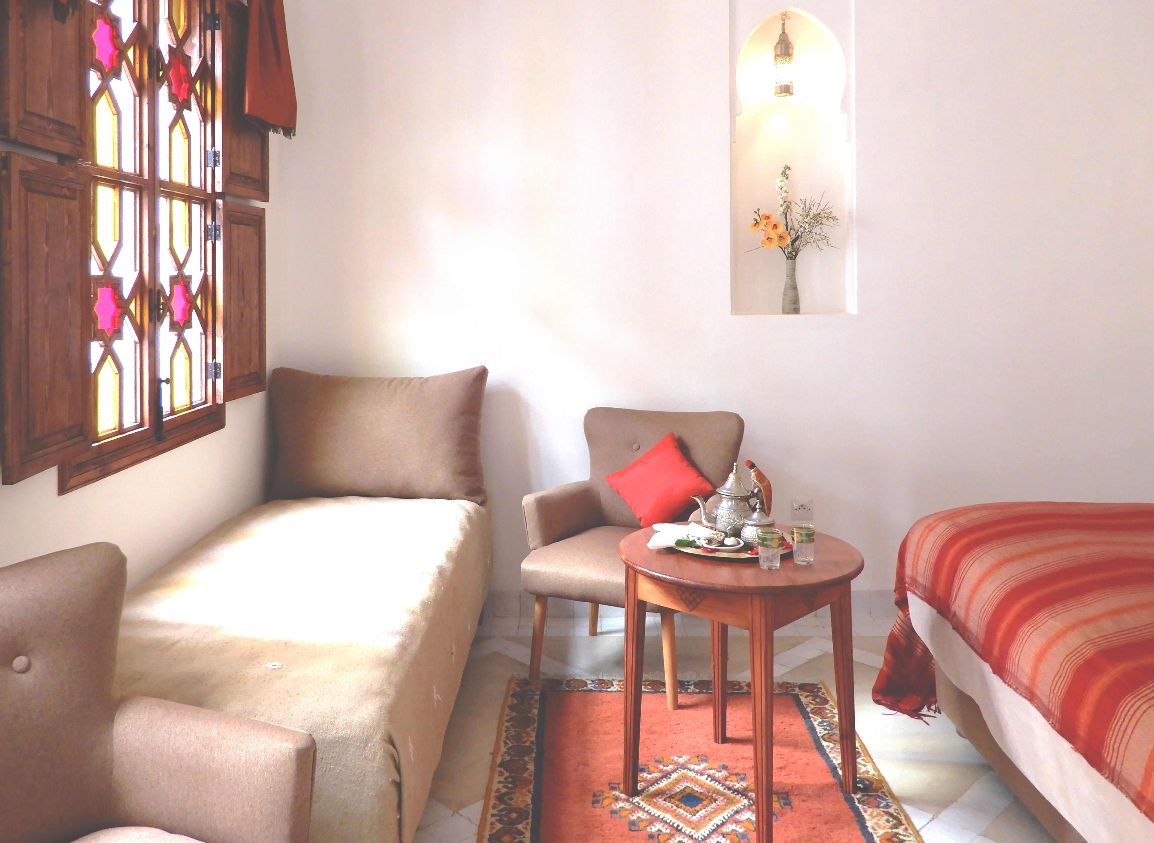 A cozy room with a bed, a chair, a small table with a teapot, and a window with decorative glass.